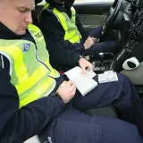 Policjanci w radiowozie spisują dokumenty. Jeden z nich trzyma w ręku dowód rejestracyjny i długopis, przygotowując się do wypisania mandatu lub grzywny.
