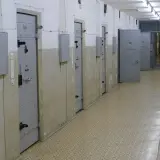 Korytarz więzienny z celami po obu stronach. Na drzwiach jednej z cel widnieje numer 106. W tle kraty i kolejne drzwi.
