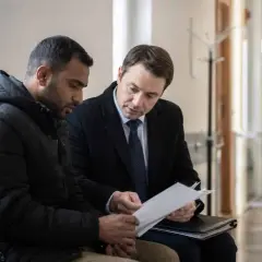 Prawnik omawia z klientem dokumenty dotyczące deportacji. W tle stoi strażnik.