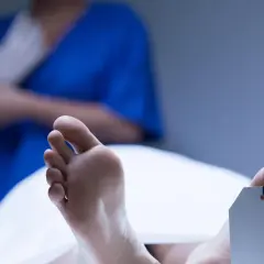 Stopy z metką identyfikacyjną, w tle osoba w niebieskiej bluzie zakłada rękawiczki. Widoczne plamy opadowe na skórze.