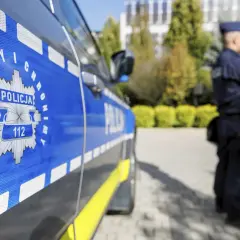 Policjant w mundurze stoi obok radiowozu z napisem "POLICJA" i numerem alarmowym 112.