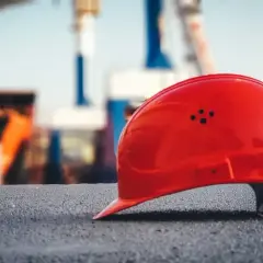 Czerwony kask leży na ziemi, symbolizując potencjalny wypadek przy pracy. W tle widać zarys budowy.