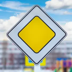 Znak droga z pierwszeństwem. Na tle błękitnego nieba z chmurami, biały romb z żółtym środkiem.