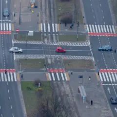 Widok z góry na skrzyżowanie z przejściami dla pieszych. Czerwony samochód wykonuje wyprzedzanie na pasach, ryzykując bezpieczeństwo.