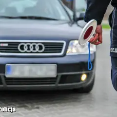 Policjant z Lubuskiej Policji zatrzymuje ruch, trzymając znak stop. Przed nim stoi ciemny samochód Audi.