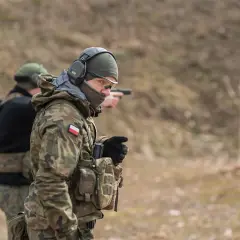 Żołnierz w mundurze maskującym, słuchawkach i okularach ochronnych celuje z pistoletu na strzelnicy.