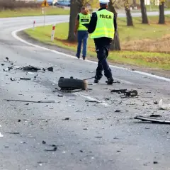 Na drodze po kolizji leżą szczątki, opona i elementy pojazdów. Policjanci w kamizelkach odblaskowych badają miejsce zdarzenia, być może szukając śladów ucieczki z miejsca kolizji.