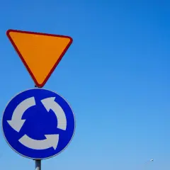Znak A7 (ustąp pierwszeństwa) i znak C12 (ruch okrężny) na tle błękitnego nieba.