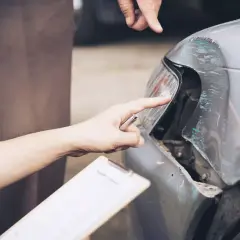 Dwóch mężczyzn ocenia uszkodzenia samochodu po wypadku. Jeden z nich, trzymający dokumenty, wskazuje na zniszczony reflektor. To zdarzenie z pewnością będzie miało wpływ na koszt naprawy, być może nawet przekroczy 288 kk.