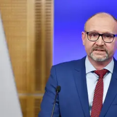 Prokurator krajowy w niebieskim garniturze, z okularami i czerwoną poszetką, przemawia przy mikrofonie. Obok widoczna flaga Polski.