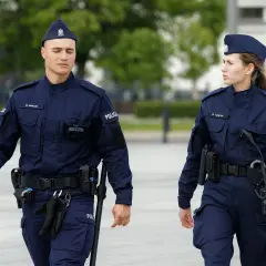 Dwóch policjantów w granatowych mundurach, z widocznymi stopniami w policji, idzie obok siebie.