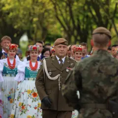 Żołnierz w brązowym capstrzyku i mundurze stoi przed grupą kobiet w tradycyjnych, kolorowych strojach ludowych.