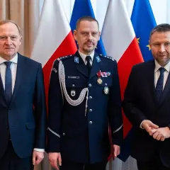 Trzech mężczyzn w garniturach i mundurze policyjnym stoi przed flagami Polski i UE.