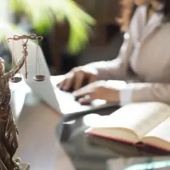 Figurka Temidy z wagą i mieczem, obok otwarta księga i osoba pracująca przy laptopie. Symbol sprawiedliwości i abolicji.