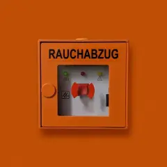 Pomarańczowa obudowa z napisem "RAUCHABZUG" i czerwoną dźwignią, kluczowy element ochrony przeciwpożarowej.