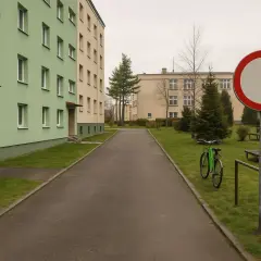Droga z zielonym rowerem i znakiem B1. Dwa bloki mieszkalne, trawnik i drzewa.