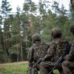 Żołnierze w mundurach kamuflażowych siedzą w lesie, patrząc w stronę drzew.