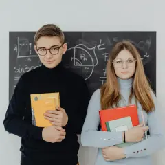 Młodociany uczeń i uczennica z książkami przed tablicą z matematycznymi wzorami.
