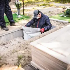 Pracownicy cmentarza przeprowadzają ekshumację, wyciągając biały worek z grobu. Obok leży drewniana trumna.