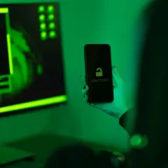 Ręka trzyma odblokowany telefon na tle monitora z zielonym kodem. Jak zrobić, by policja nie namierzyła telefonu?