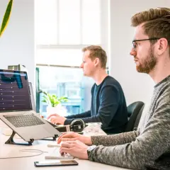 Dwóch programistów pracuje przy komputerach. Jeden z nich ma okulary, co może sugerować refundację okularów przez pracodawcę.