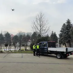 Dron policyjny w powietrzu, obok radiowozu i policjantów.