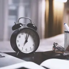 Budzik pokazuje prawie dwunastą, symbolizując pracę w nocy i dodatek za nią. Obok laptop i przybory biurowe.