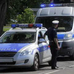 Policjant stoi obok radiowozu i furgonetki policyjnej z włączonymi sygnałami świetlnymi.