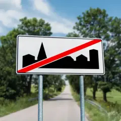 Znak D42: koniec obszaru zabudowany. Droga prowadzi przez zielone pola i lasy, a niebo jest błękitne z chmurami.