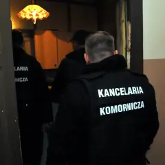 Trzech mężczyzn w czarnych kamizelkach z napisem "KANCELARIA KOMORNICZA" wchodzi do pomieszczenia. Prawdopodobnie to postępowanie egzekucyjne.
