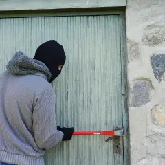 Osoba w masce próbuje wyważyć drzwi łomem. To klasyczny przykład kradzieży z włamaniem.