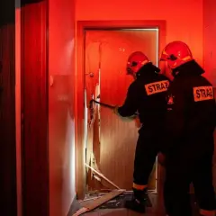 Strażacy wyważają drzwi, gdy kobieta patrzy z niepokojem. Sytuacja, kiedy policja może wyważyć drzwi, by ratować życie lub zapobiec przestępstwu.