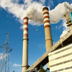 Dwie wysokie, prążkowane kominy elektrowni emitują dym na tle błękitnego nieba. Obok widać linie energetyczne i budynki przemysłowe, tworzące obraz kluczowej infrastruktury.