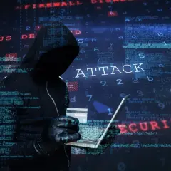 Sylwetka hakera w kapturze, trzymająca laptopa i kartę kredytową. Na tle widać kod i napisy "ATTACK", "VIRUS", "MALWARE", "SECURITY", sugerujące atak hakerski.