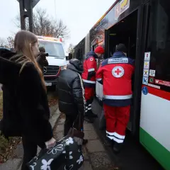 Osoby z bagażami wsiadają do autobusu z pomocą humanitarną. W tle biała furgonetka, przypominająca pojazd z lotniska, gdzie mógł wystąpić alarm.