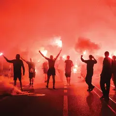 Tłum ludzi, wśród nich chuligan, idzie ulicą w gęstym, czerwonym dymie z rac.