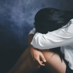 Kobieta w białej koszuli skulona, przeżywająca traumę po gwałcie. Ciemne tło podkreśla jej cierpienie.