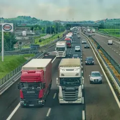 Ciężarówki na autostradzie, jedne wymijają, inne omijają. W tle znak Esso i zielone wzgórza.