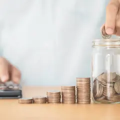 Osoba wrzuca monetę do słoika, obok rosnące stosy monet i kalkulator. To jakby finansowy dyżur, budowanie oszczędności.