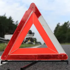 Trójkąt ostrzegawczy, część obowiązkowego wyposażenia samochodu, stoi na poboczu drogi. W tle widać przewrócony samochód ciężarowy.
