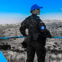 Żołnierz w mundurze z naszywką ONZ, w niebieskiej czapce, stoi na tle krajobrazu. Może to być kapelan na misji.