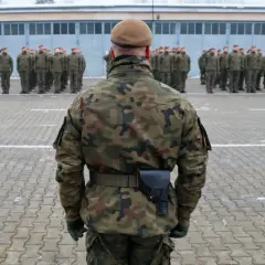 Żołnierz w mundurze moro i beretcie stoi na placu apelowym przed szeregiem innych żołnierzy.