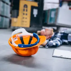 Pomarańczowy kask leży obok nieprzytomnego pracownika. To smutny obraz wypadku w pracy.