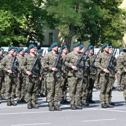 Żołnierze w mundurach i zielonych beretach, z karabinami w rękach, stoją w szeregu. W tle zielona kula z napisem AWL.