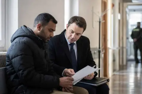 Prawnik omawia z klientem dokumenty dotyczące deportacji. W tle stoi strażnik.