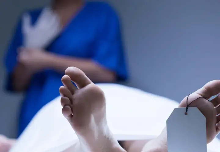 Stopy z metką identyfikacyjną, w tle osoba w niebieskiej bluzie zakłada rękawiczki. Widoczne plamy opadowe na skórze.