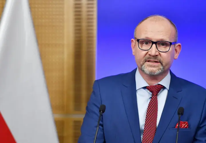 Prokurator krajowy w niebieskim garniturze, z okularami i czerwoną poszetką, przemawia przy mikrofonie. Obok widoczna flaga Polski.