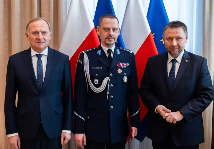 Trzech mężczyzn w garniturach i mundurze policyjnym stoi przed flagami Polski i UE.