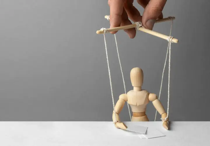 Drewniany ludzik-marionetka, sterowany przez ludzką dłoń, symbolizuje podżeganie i manipulację.