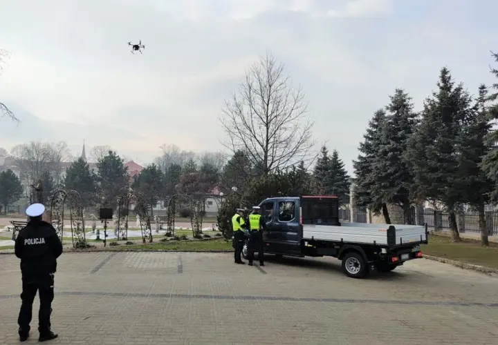 Dron policyjny w powietrzu, obok radiowozu i policjantów.